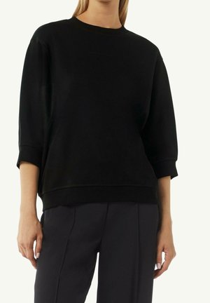 Mujer con sudadera negra de manga corta y suelta y pantalones negros a medida, de pie con los brazos relajados a los lados.