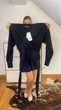 Ženska drži črno dolg rokav ovitka pulover z etiketami, stoji na rjavi in belih kravjih kožah v prostoru z lesenim podom.