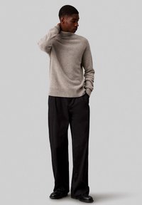 Maglione color grigio chiaro a dolcevita con polsini e orlo inferiore a coste, abbinato a pantaloni neri a gamba larga e scarpe nere, su sfondo neutro.