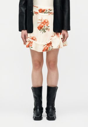 Persona con falda cremosa con volantes y estampado floral, chaqueta de cuero negra y botas negras hasta la mitad de la pantorrilla, de pie contra un fondo blanco liso.