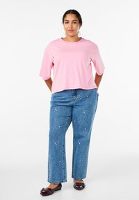 Lyserød cropped t-shirt med korte ærmer, parret med blå denim jeans med blomsterbroderi. Modellen har maroon flade sko på.