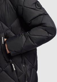 Veste matelassée noire avec un fini brillant ; elle comprend une poche zippée et un écusson rond avec un logo. Le tissu semble lisse avec une légère brillance.