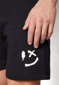 Shorts noirs en tissu doux, avec un imprimé graphique blanc représentant un visage souriant, une éclaboussure et un X. Gros plan avec une main touchant le tissu.