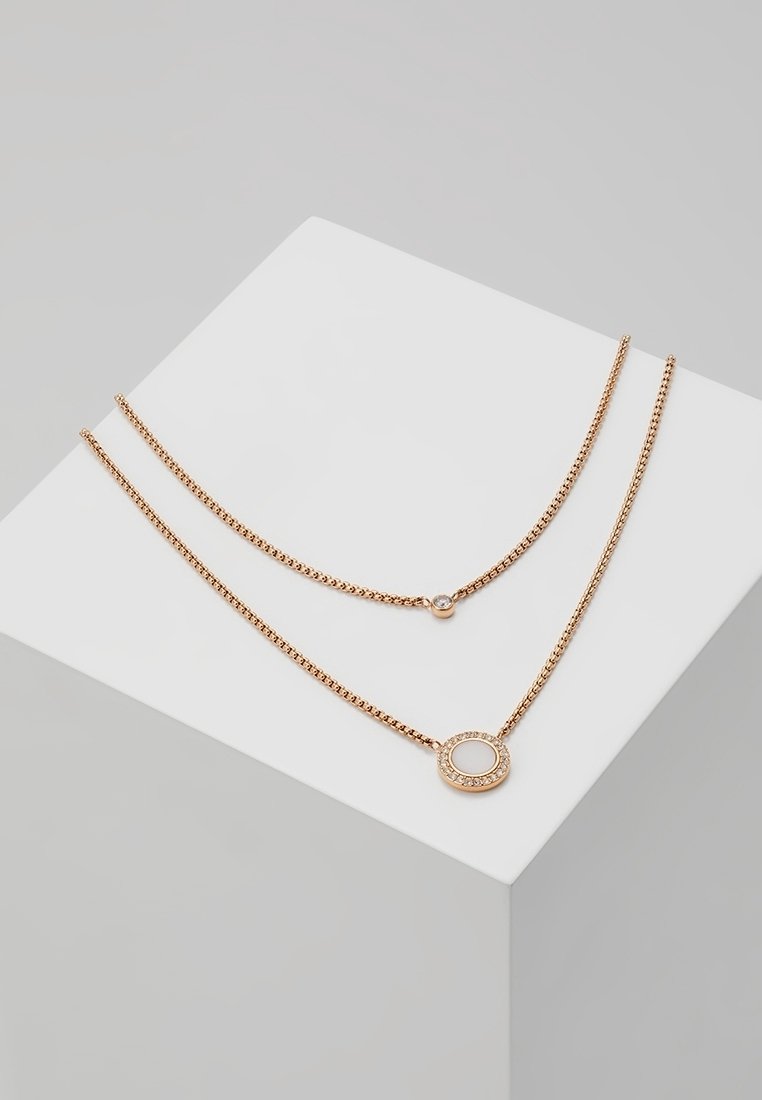 Deux délicats colliers en or avec de petits pendentifs circulaires présentés sur un cube blanc sur fond gris.