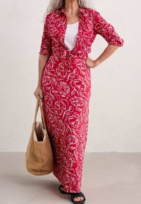 Vestido maxi rojo con flores y dibujos lineales blancos, mangas cortas y delantero anudado. Lleva sandalias negras y sostiene un bolso de yute beige.
