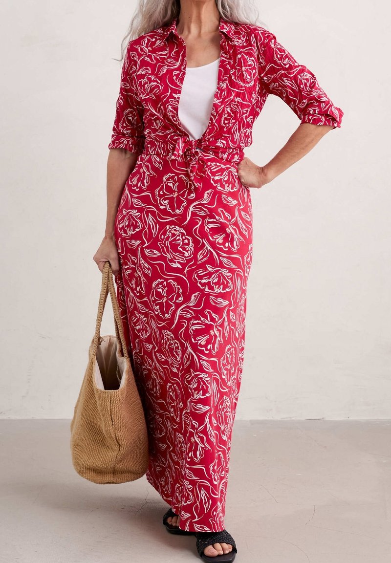 Vestido maxi rojo con flores y dibujos lineales blancos, mangas cortas y delantero anudado. Lleva sandalias negras y sostiene un bolso de yute beige.