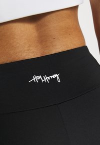 Černé legíny s vysokým pasem mají hladký povrch a bílé logo "Hey Honey" na pasu, což podtrhuje jejich přiléhavý střih.