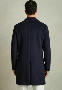 Mørkeblå uldfrakke med lige snit, der har en enkelt rygslids og stramme ærmer. Glat tekstur, minimalistisk design og ingen synlige metaldele.