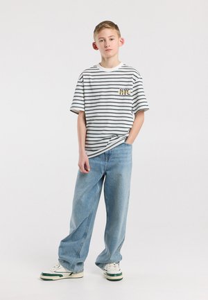 Tienerjongen staand met één hand in zijn zak, draagt een wit met zwart gestreept NYC T-shirt, losse blauwe spijkerbroek en witte sneakers.