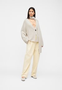 Γυναίκα που στέκεται φορώντας μπεζ παντελόνι με φαρδύ πόδι, ανοιχτό γκρι ζακέτα με βαθύ V λαιμόκοψη και ένα κουμπί, ταιριαστό μαντήλι και κολιέ με μαργαριτάρια.