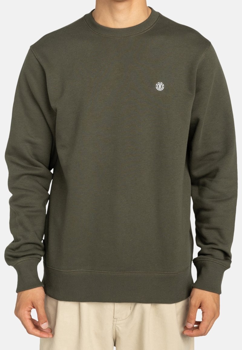 Homme portant un sweat-shirt vert olive à col rond avec un petit logo blanc circulaire sur la poitrine gauche et un pantalon beige, debout, bras détendus.