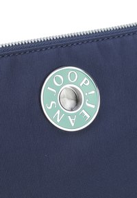 Borsa in nylon blu marino con zip argentata. Presenta un accento rotondo in verde menta con testo inciso, posizionato su uno sfondo di tessuto testurizzato.