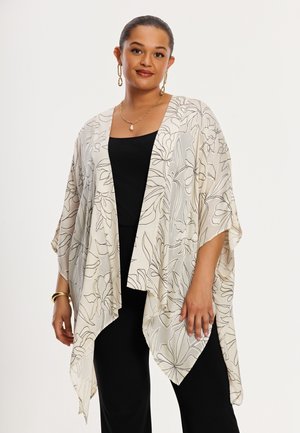 Vrouw die een witte kimono-achtige cardigan met bloemenprint draagt over een zwarte top en broek, geaccentueerd met gouden oorbellen, ketting en armband.