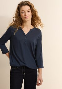 Marineblaues Langarmshirt mit Henley-Ausschnitt, drei Knöpfen und lockerer Passform, aus weichem, glattem Material. Getragen mit dunklen Jeans.