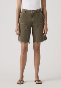 Γυναίκα που φοράει σορτς μήκους μέχρι το γόνατο σε ελιά πράσινο με τσέπες στο μπροστινό μέρος και ακατέργαστο τελείωμα, συνδυασμένα με μαύρα σανδάλια τύπου σαγιονάρα.