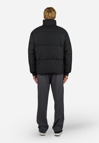 Geacă puffer neagră cu guler înalt, având o siluetă pătrățoasă, secțiuni matlasate și manșete elastice, asortată cu pantaloni gri.