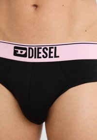 Black cotton briefs featuring a pink elastic waistband with black "DIESEL" logo. Smooth texture and fitted design.