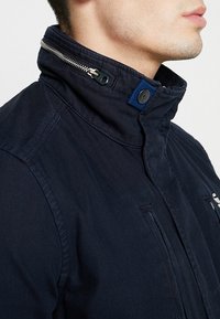 Marinblå jacka med hög krage, dragkedjedetaljer och knappaccenter. Strukturerad tyg, med två fickor med dragkedja på framsidan.