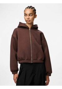 Felpa con zip marrone dal fit comodo, con polsini e orlo a costine. Presenta un cappuccio foderato, una texture liscia e un design minimalista.