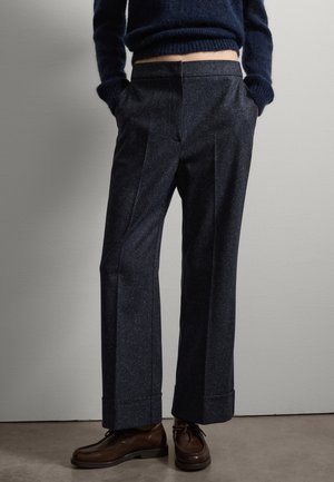 Pantalons en laine bleu marine foncé avec une finition texturée, jambes larges et revers, associés à des chaussures en cuir marron. Matière douce et lisse.