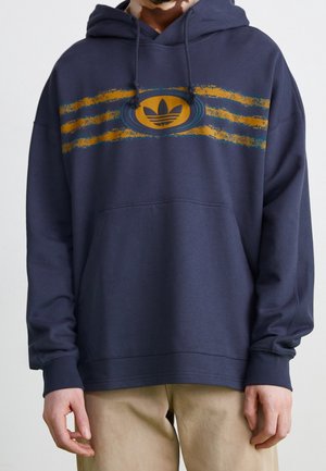 Laivastonsininen huppari, jossa keltaisia ja sinisiä raitoja sekä pyöreä logo rinnassa, päällä henkilö, jolla on beiget housut ja kädet rentoina sivuilla.