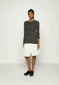 Camisola preta de mangas longas com padrão de bolinhas brancas, combinada com uma saia branca solta. O modelo usa tamancos pretos e segura uma bolsa pequena.