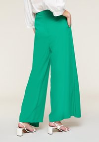 Vida byxor i klar grön tyg, med hög midja och sidofickor, ihopparade med vita blockklackade sandaler.