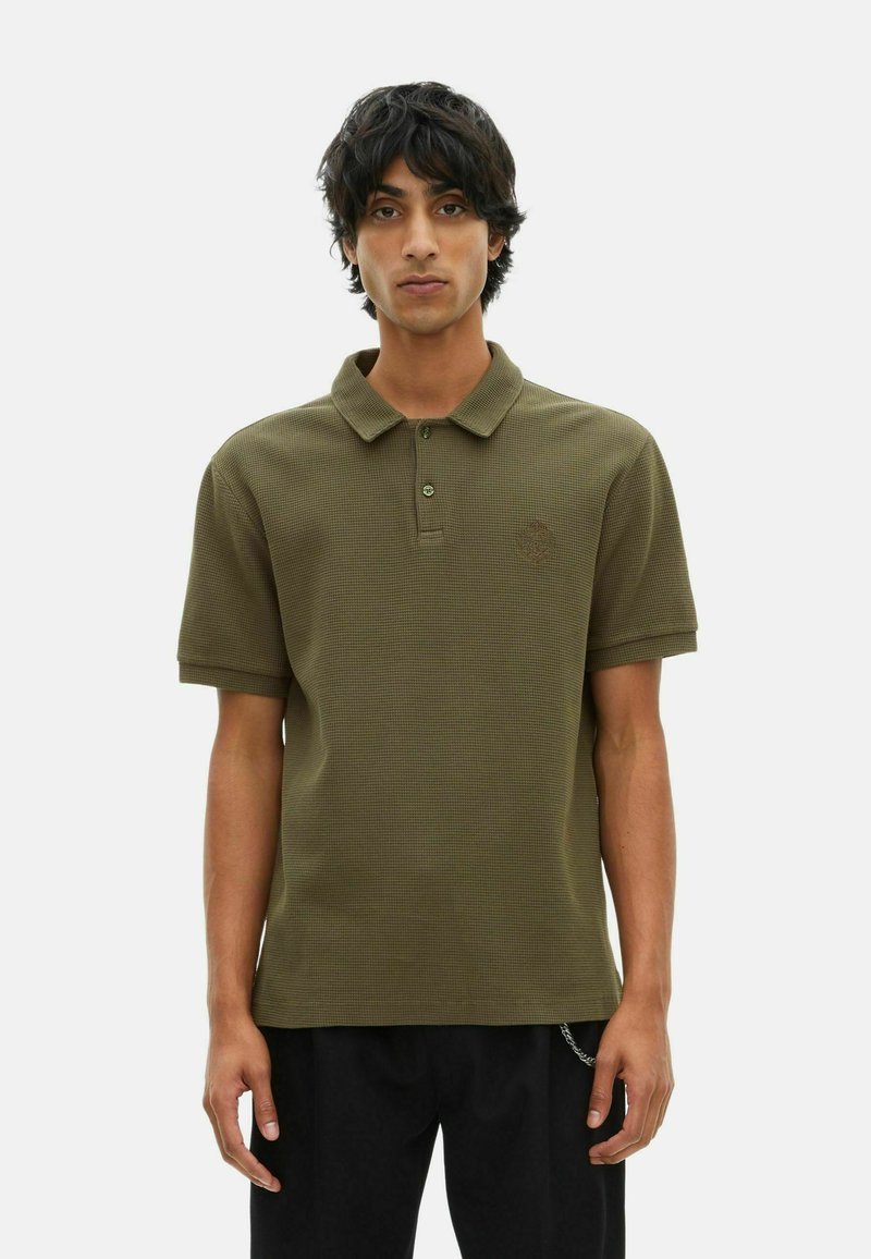 Giovane uomo che indossa una polo a maniche corte texturizzata di colore verde oliva e pantaloni neri, in piedi contro uno sfondo chiaro e semplice.