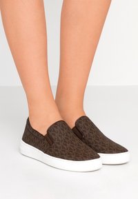 Chaussures marron à enfiler en tissu texturé, ornées d'un motif géométrique, avec des accents élastiques sur les côtés et des semelles en caoutchouc blanc.