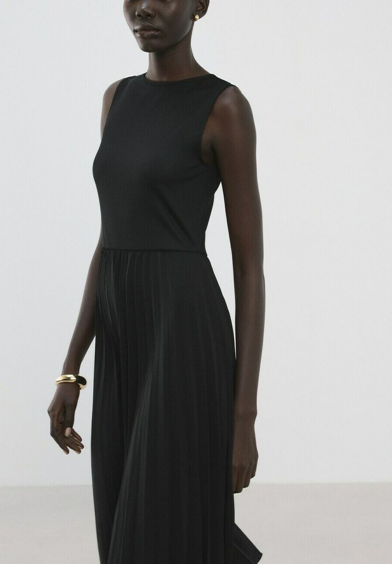 Femme portant une robe noire plissée sans manches et un bracelet en or, debout devant un fond clair uni.