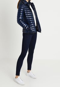 Padded navy jacket with a shiny finish, featuring a zippered front, stand collar, and side pockets, worn over dark jeans and white sneakers.
