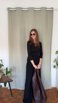 Μαύρο τοπ με μακριά μανίκια και μαύρο παντελόνι ψηλόμεσο, κρατώντας μια καφέ δερμάτινη ζακέτα. Ανοιχτοπράσινη κουρτίνα στο φόντο. Ξύλινο πάτωμα.