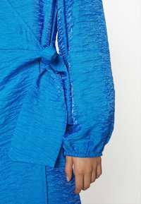 Primer plano de la mano de una persona junto a un vestido de tela texturizada azul brillante con mangas fruncidas y un detalle de cintura atada.