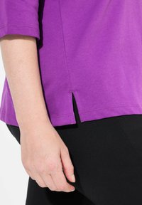 Lila Baumwoll-T-Shirt mit kurzen Ärmeln, ausgestattet mit einem gesplitteten Saumdesign, getragen mit schwarzen Hosen. Glatte Textur und lässige Passform.