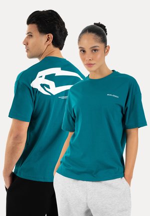 Homme et femme portant des t-shirts bleu sarcelle, l'homme montre un graphique de flèche blanche dans le dos, la femme a un petit logo à l'avant.