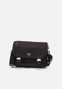 Sort rektangulær messenger-taske med justerbar skulderrem, frontklap, spænder i sølvfarve og trekantet Guess-logo-badge.