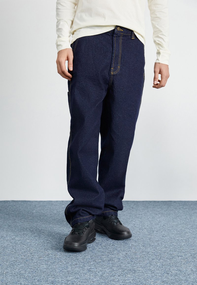 Blå denimbukser med en løs pasform, kontrasterende gul syning og sidelommer. Båret med sorte sneakers på et struktureret blåt tæppe.