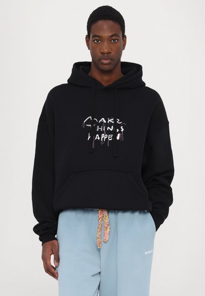 Black hoodie with a front pocket, featuring multicolored embroidered text “Make Things Happen.” Paired with light blue sweatpants with a textured drawstring.