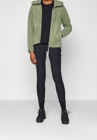 Groene fleecetrui met hoge kraag, ritsluiting en logo op de borst, gecombineerd met zwarte slim-fit broek en zwarte wandelschoenen.
