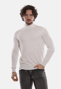 Maglione bianco attillato a collo alto con una superficie liscia, maniche lunghe e polsini a coste. Indossato con jeans in denim scuro.