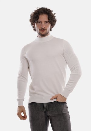 Maglione bianco attillato a collo alto con una superficie liscia, maniche lunghe e polsini a coste. Indossato con jeans in denim scuro.