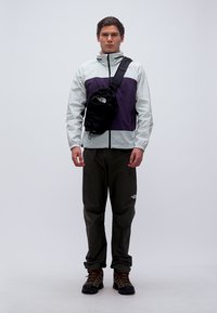 Άνδρας με ελαφρύ μπουφάν, μαύρη τσάντα ώμου, παντελόνι πεζοπορίας και παπούτσια πεζοπορίας, στέκεται μπροστά σε απλό φόντο.