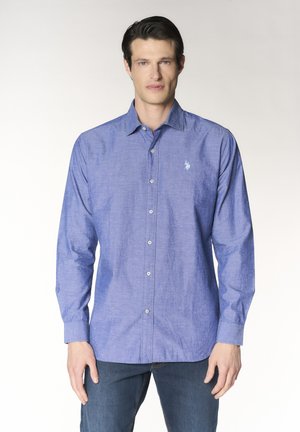 Uomo che indossa una camicia blu a maniche lunghe con un piccolo logo bianco sul petto, abbinata a jeans blu, in piedi contro uno sfondo chiaro.