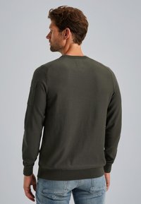 Maglione verde scuro con scollo rotondo e maniche lunghe. Presenta una finitura testurizzata e una toppa sulla manica sinistra. Design liscio e aderente.