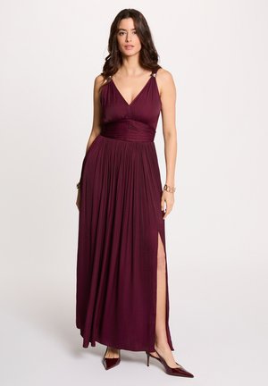 Donna con abito lungo color prugna senza maniche, con pieghe, spacco laterale, dettagli in cinturini dorati e tacchi a punta abbinati, in piedi davanti a uno sfondo semplice.