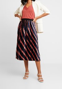 Kvinna klädd i en vit kavaj, rostfärgad V-ringad topp, plisserad marinblå kjol med diagonala röda mönster och vita sandaler med remmar, står med händerna på höfterna.