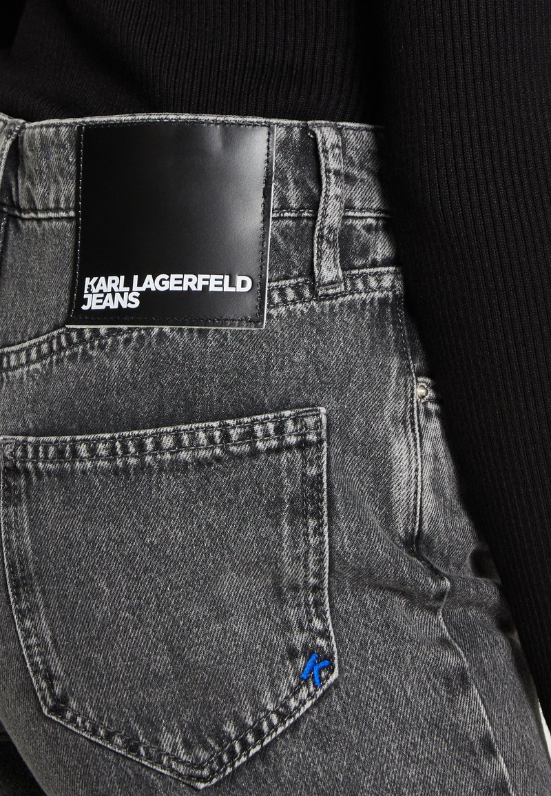 Un jean en denim gris présente une étiquette en cuir noir avec "KARL LAGERFELD JEANS" embossé en blanc. Un accent bleu sur la poche arrière ajoute une touche de détail.