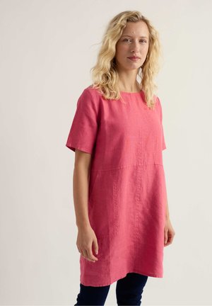 Hullámos hajú szőke nő áll rövid ujjú rózsaszín lenruhában és sötét nadrágban, egyszínű világos háttér előtt.