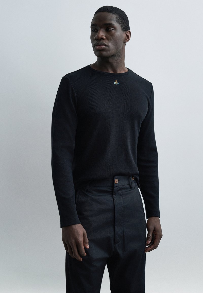 Άνδρας που φοράει μαύρο μακρυμάνικο πουκάμισο με μικρό πολύχρωμο έμβλημα στο στήθος και σκούρο παντελόνι, στέκεται μπροστά σε απλό ανοιχτόφωτο φόντο.