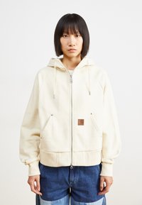 Cream zip-up hoodie with a drawstring hood, front pockets, and a brown logo patch, featuring a relaxed fit and ribbed cuffs and hem.